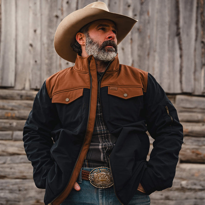 Man wearing a black and coyote brown Augusta jacket, cowboy hat, and denim jeans, standing against a rustic wooden backdrop.