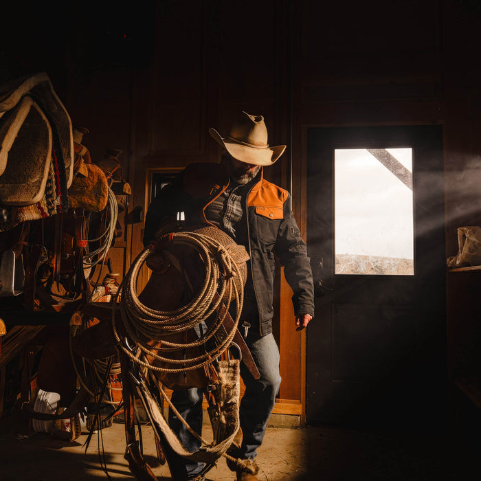 Man wearing a black and coyote AUGUSTA JACKET, holding a rope in a rustic barn setting.