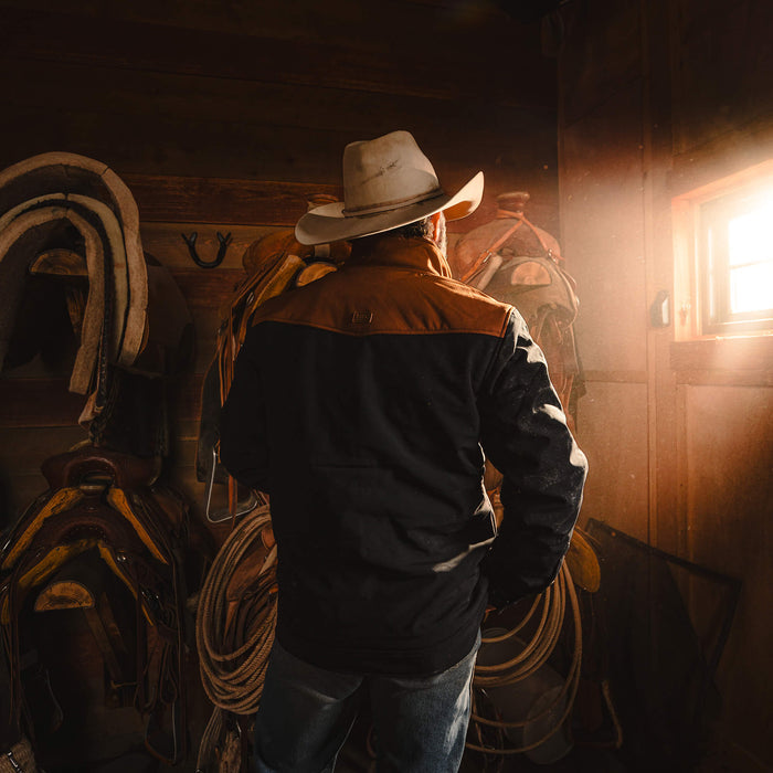 A man wearing a black and coyote AUGUSTA JACKET stands in a barn, surrounded by saddles and ropes, with sunlight streaming in.