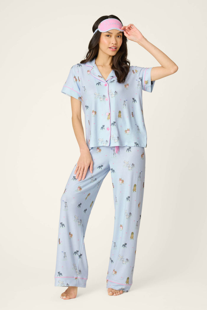 A woman models the P.J. Salvage Bark Park Fetch & Frolic 3-Piece PJ Set—light blue dog-print pajamas with matching pants and a pink sleep mask—standing barefoot, perfect for cozy women's sleepwear.