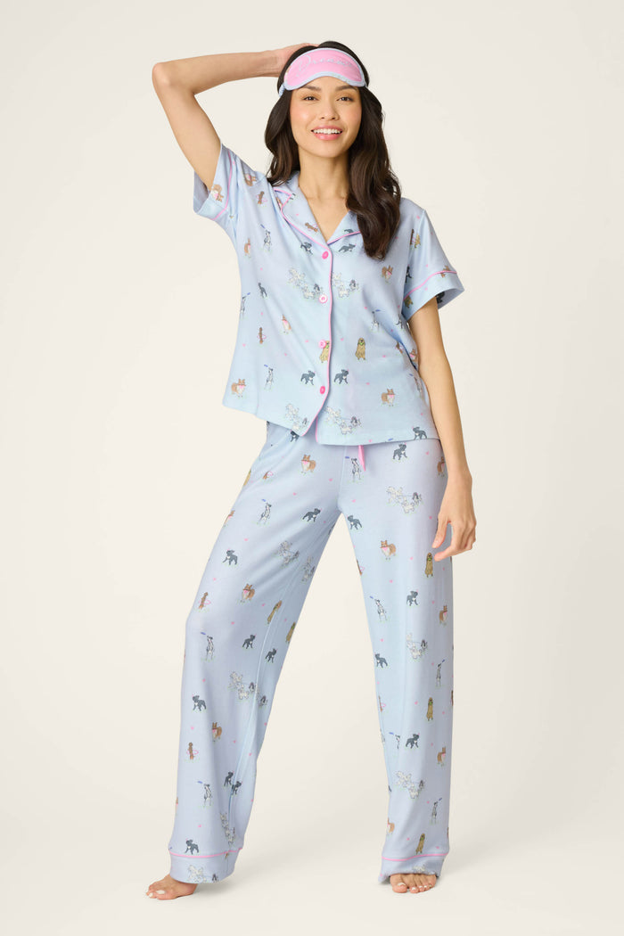A woman barefoot and smiling stands against a plain background, wearing the P.J. Salvage Bark Park Fetch & Frolic 3-Piece PJ Set—colorful dog lover pajamas with pink trim—while holding a pink sleep mask on her head.