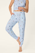 A person poses barefoot wearing P.J. Salvage Bark Park Fetch & Frolic Banded Pants—a light blue pajama with a drawstring waist and colorful animal and character prints—on a plain background.