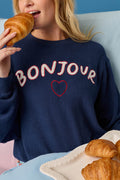 A woman in a navy blue P.J. Salvage Bonjour Babe Boucle Bonjour Sweater holds a croissant near her mouth. A plate with more croissants sits in front of her against a light blue background.
