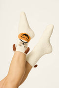 Two crossed legs wearing fuzzy white "Boyfriend Boxer Trick-or-Treating Dog Socks" by P.J. Salvage, each featuring an adorable dog face with orange pumpkin accents on the front and small brown ear and tail details. The background is plain and light-colored.