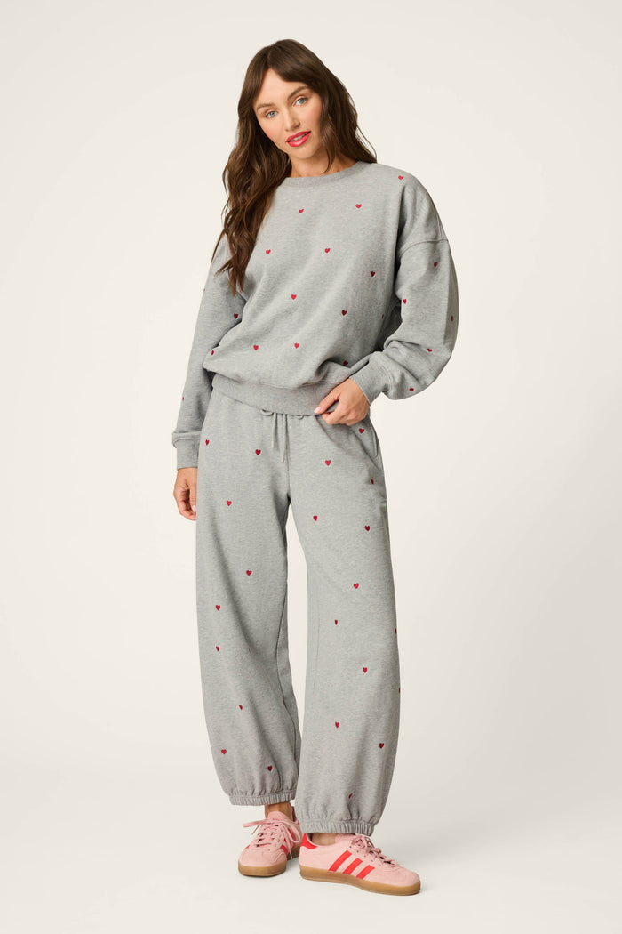 A woman stands smiling against a plain white background, dressed in the P.J. Salvage C'est La Vie Embroidered Hearts Frankie Sweatshirt—a loose-fitting gray sweatsuit adorned with small red heart patterns—and paired with pink Adidas sneakers.