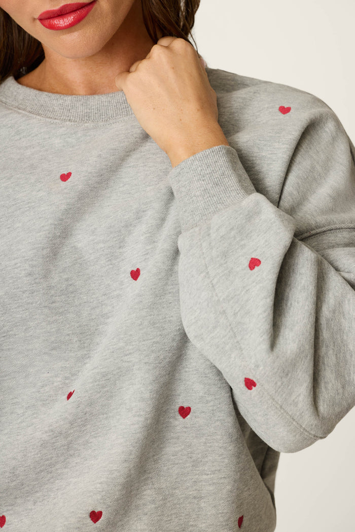 A person wearing the P.J. Salvage C'est La Vie Embroidered Hearts Frankie Sweatshirt, featuring a light gray fabric with small red heart designs, holds their fist near their chin. Only the lower part of their face with red lipstick and upper torso are visible.