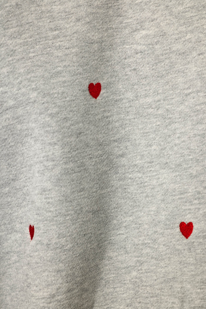 Close-up of a light heather gray sweatshirt style fabric with small red hearts embroidered into the fabric and scattered across it, widely spaced apart.