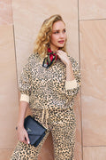 A woman in the C'est La Vie Leopard Print Frankie Sweater by P.J. Salvage poses against a beige stone wall, holding a black Prada clutch and wearing a red patterned scarf around her neck. She has wavy blonde hair and looks off to the side, smiling slightly.