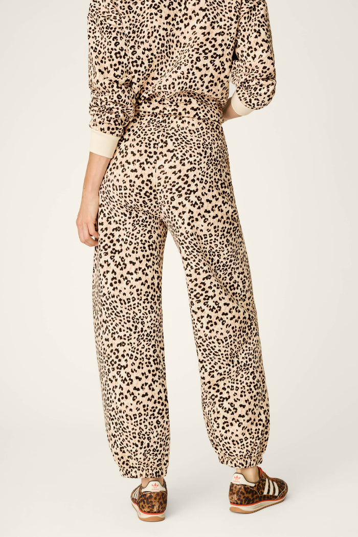 A person stands facing away, wearing the P.J. Salvage C'est La Vie Leopard Print Good Hang Jogger Pants in a beige and black leopard print set, paired with brown sandals. The background is plain and light-colored.