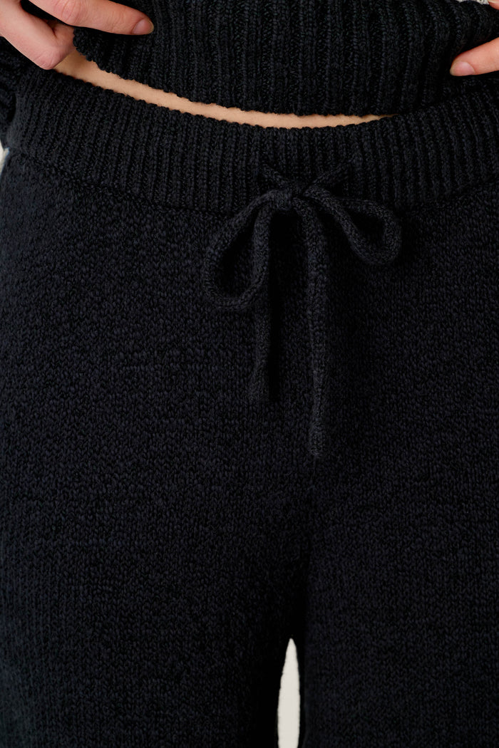 Close-up of a person wearing the C'est La Vie Suki Jogger Pants by P.J. Salvage, featuring a black textured knit fabric and a drawstring waist. The person's hands rest on their waist, with the bottom edge of a coordinating knit top also visible.