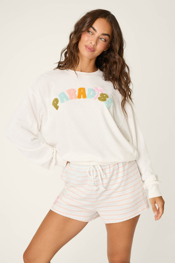 A woman models the P.J. Salvage Cheeky Paradise Sweatshirt, featuring colorful "PARADISE" lettering, paired with light striped shorts and posing with one hand on her hip, gazing to the side.
