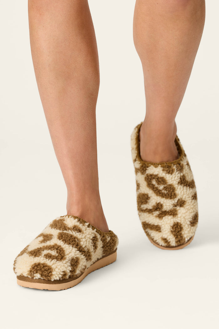 A person wearing P.J. Salvage Cheetah Bonita Dreams cheetah print fuzzy slippers is standing on a light-colored surface.