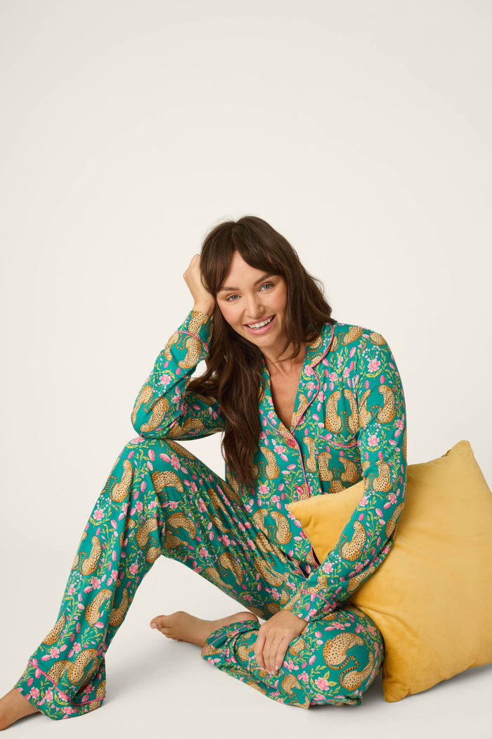 Wearing the P.J. Salvage Cheetah Bonita Dreams Cheetah Print Long Sleeve PJ Set, a woman sits on the floor, smiling and leaning on a yellow pillow against a plain background.