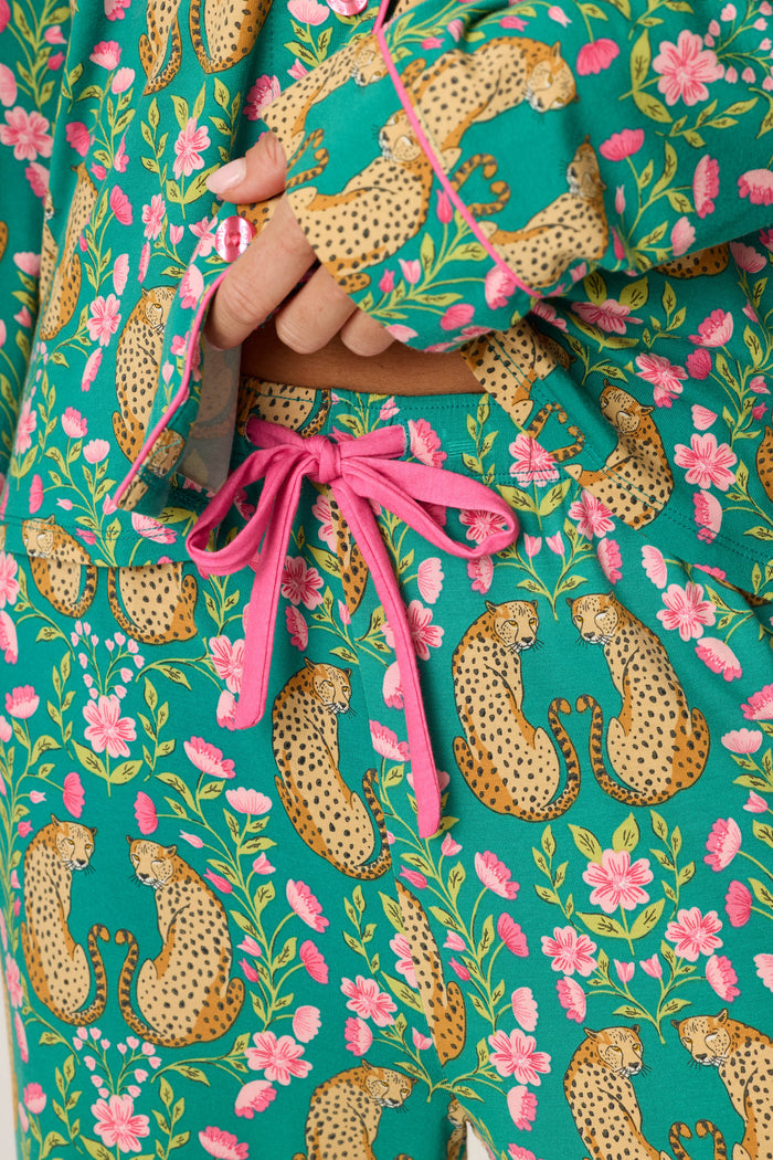 A close-up of a person wearing the P.J. Salvage Cheetah Bonita Dreams Cheetah Print Long Sleeve PJ Set shows teal pajamas with a pink drawstring, adorned with cheetahs and pink flowers. The person’s hand rests near the waistband.