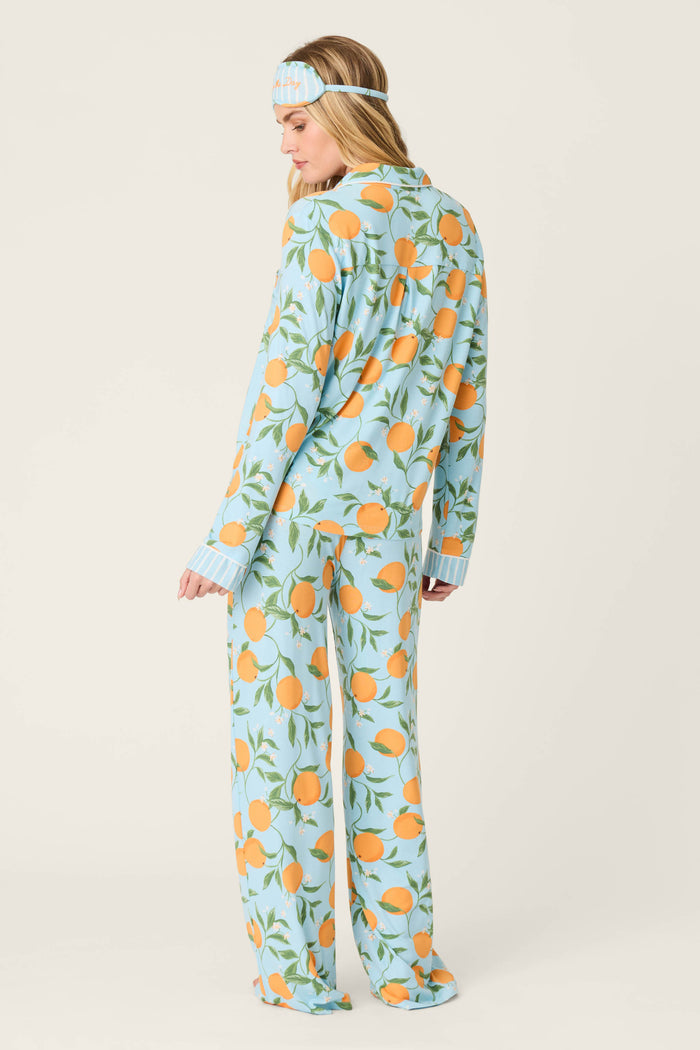A woman stands turned slightly away, wearing the Citrus Dreams Morning Squeeze Long 3-Piece PJ Set by P.J. Salvage, featuring a blue top and citrus-print sleep mask with oranges and green leaves against a plain, light backdrop.