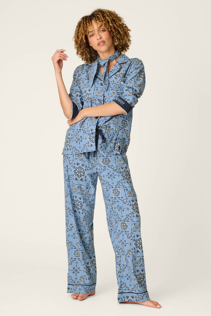 A person with curly hair wears the P.J. Salvage Classic Paisley Bandana Print 3-Piece Long Sleeve Flannel PJ Set, featuring a loose-fitting, blue paisley pattern. Standing barefoot against a light background, they have one arm raised while the other rests relaxed by their side.