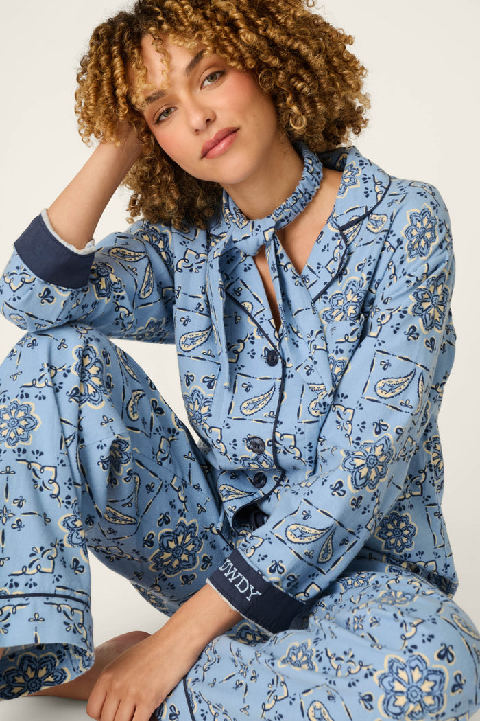 A woman with curly hair sits barefoot wearing the P.J. Salvage Classic Paisley Bandana Print 3-Piece Long Sleeve Flannel PJ Set. She has one arm resting on her knee and a soft smile, with IMPY embroidered on the cuff. The background is plain and light-colored.
