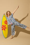 A woman in the Cotton Jersey Midnight Garden Cropped PJ Set by P.J. Salvage stands on one leg, smiling and balancing with arms outstretched as she holds a yellow surfboard with large red flowers on a beige background.