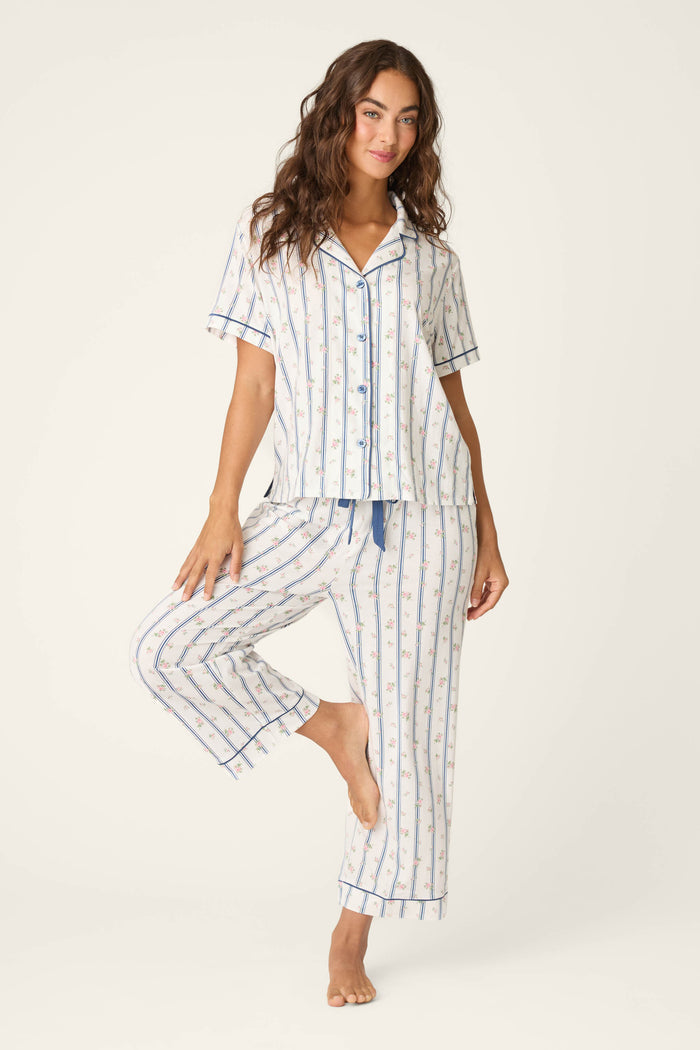 A woman with long brown hair poses barefoot and smiling, wearing the P.J. Salvage Cotton Jersey Stripes & Blooms Cropped PJ Set, a matching striped pajama with blue trim and a button-up short-sleeve top. She stands on one leg with the other slightly raised.
