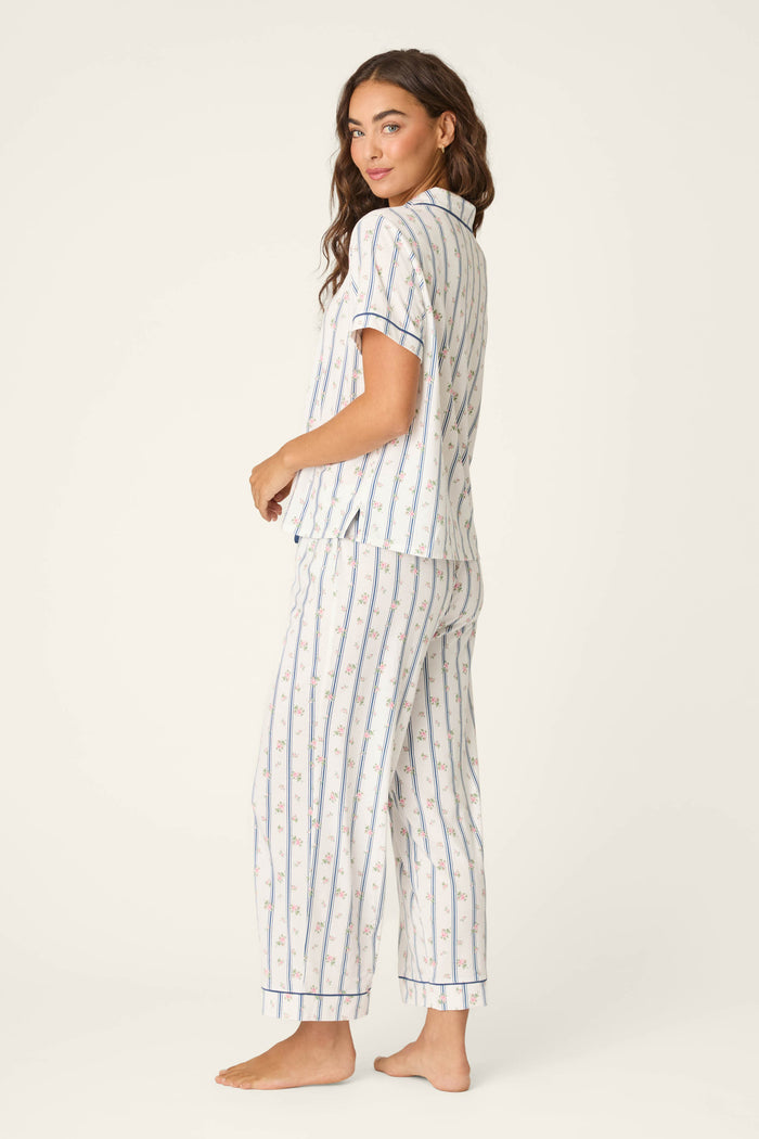 A woman stands barefoot, looking over her shoulder, wearing the P.J. Salvage Cotton Jersey Stripes & Blooms Cropped PJ Set featuring vertical blue stripes and floral patterns on a light background, with a short-sleeve top and ankle-length pants.