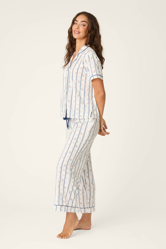 A woman with long brown hair poses barefoot, smiling gently in the P.J. Salvage Cotton Jersey Stripes & Blooms Cropped PJ Set—featuring a white and blue striped short-sleeve button-up top and loose pants—against a plain light background.