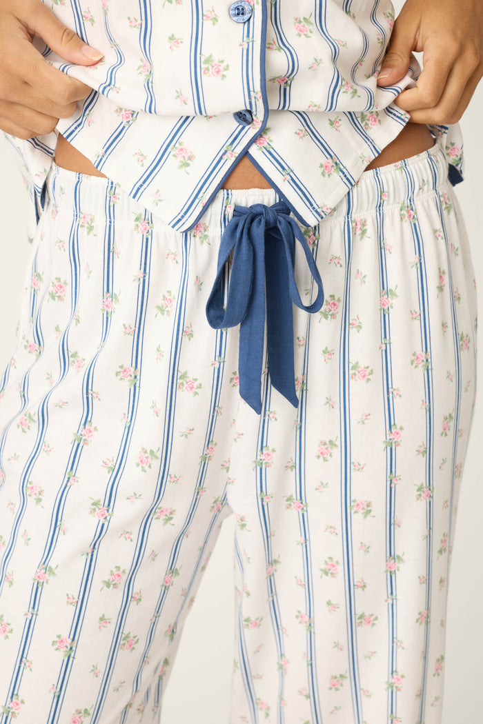 A person wears the P.J. Salvage Cotton Jersey Stripes & Blooms Cropped PJ Set, featuring white pajamas with blue stripes and pink floral accents. The pants have a blue drawstring, the shirt is slightly unbuttoned, and hands rest on the waist.
