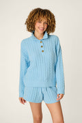 A person wearing the P.J. Salvage Cozy Chenille Cable Knit Cloud Blue Pullover, smiling while standing against a plain, light background.