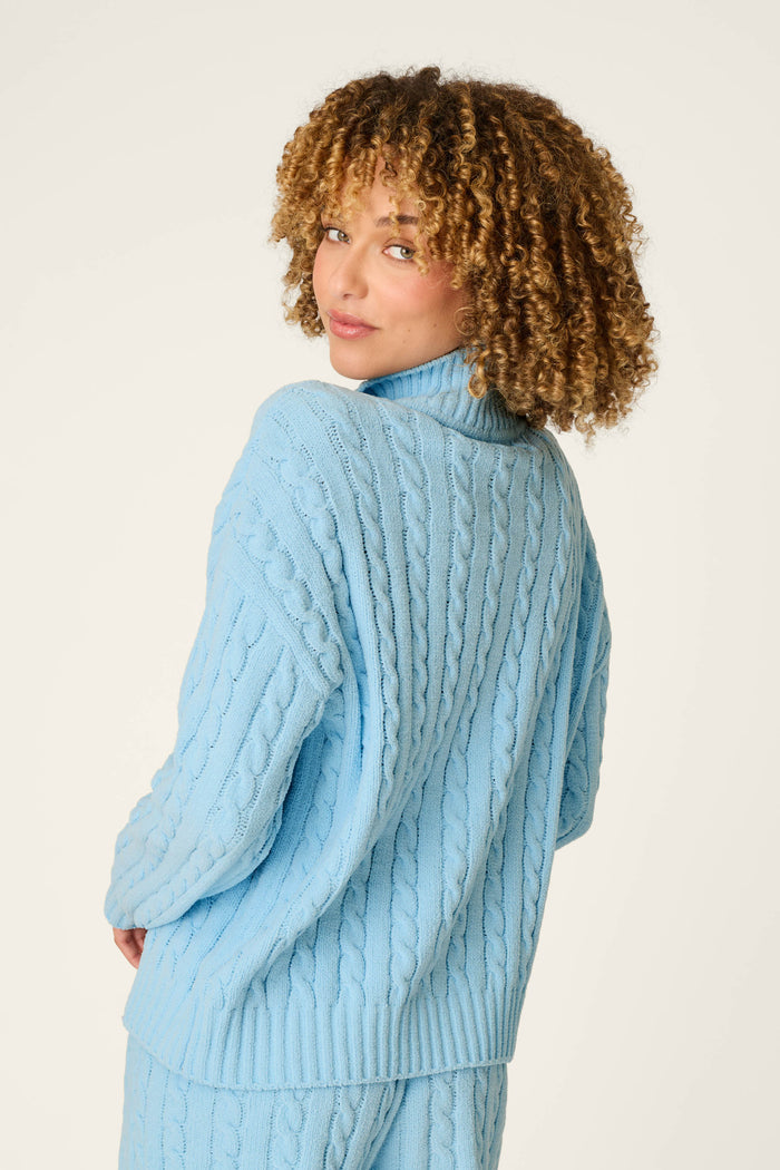 Wearing the Cozy Chenille Cable Knit Cloud Blue Pullover by P.J. Salvage, a person with curly hair looks over their shoulder at the camera against a plain light background.