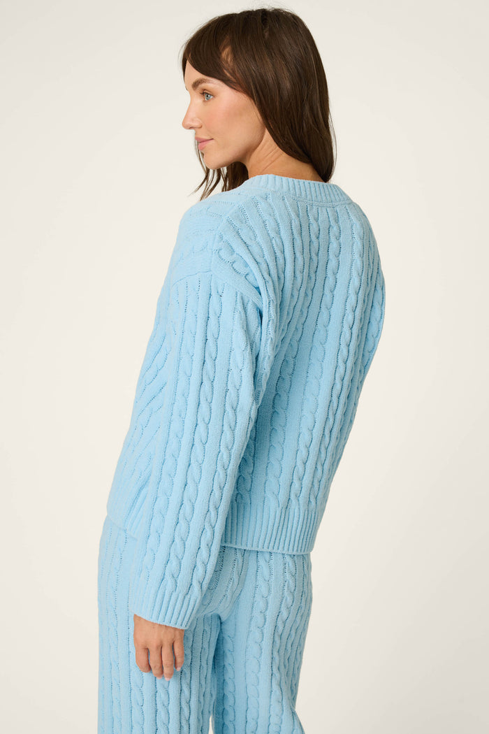 A woman with brown hair wears the P.J. Salvage Cozy Chenille Cable Knit Cloud Blue V-Neck Sweater, paired with matching pants, as she stands against a plain light background looking to the side.
