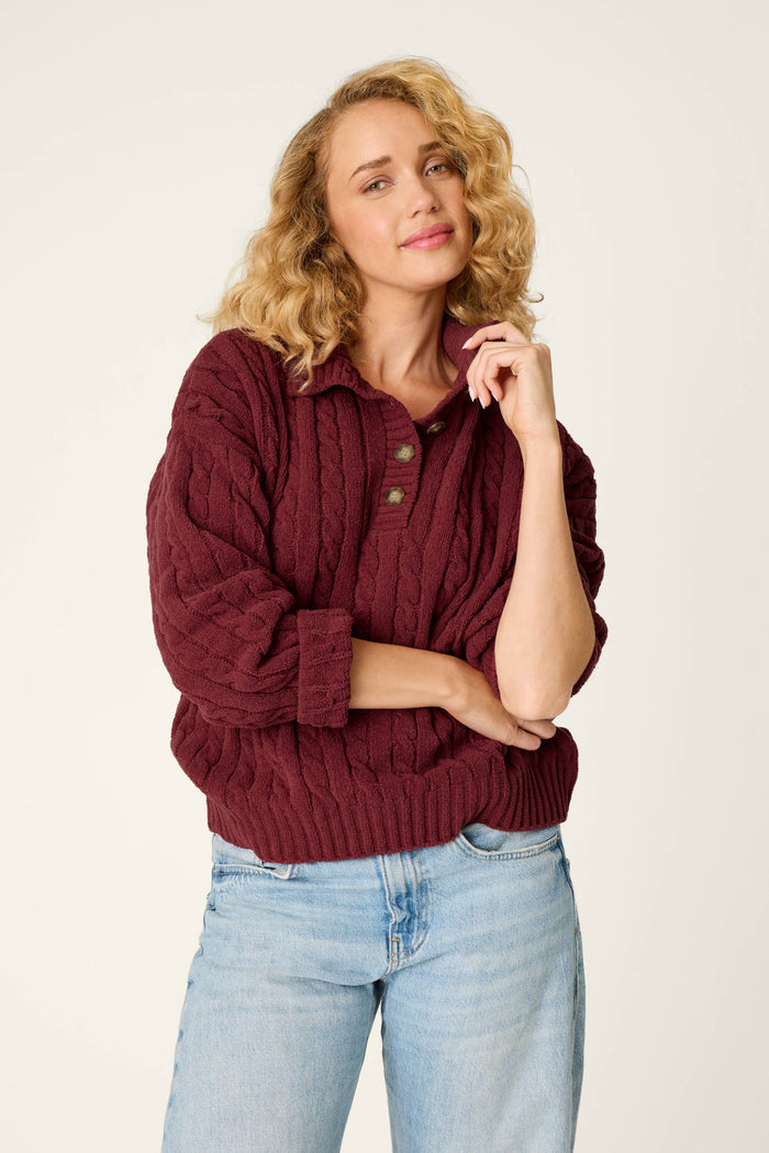 A woman with curly blonde hair wears the P.J. Salvage Cozy Chenille Cable Knit Dark Wine Pullover and light blue jeans, standing against a plain light background with a relaxed expression and one hand touching her face.