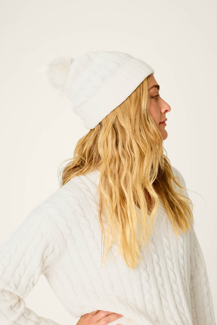 A woman wearing a Cozy Chenille Cable Knit Ivory Beanie with Pompom by P.J. Salvage stands sideways with her hand on her hip, looking off to the right against a plain light background.