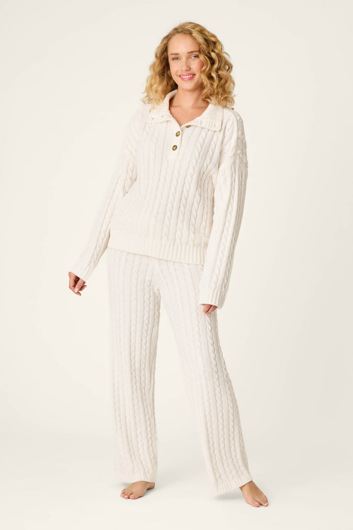 A woman stands barefoot and smiles against a plain light background, wearing the P.J. Salvage Cozy Chenille Cable Knit Ivory Pullover Lounge Set, a matching cream-toned ensemble with cable patterns.
