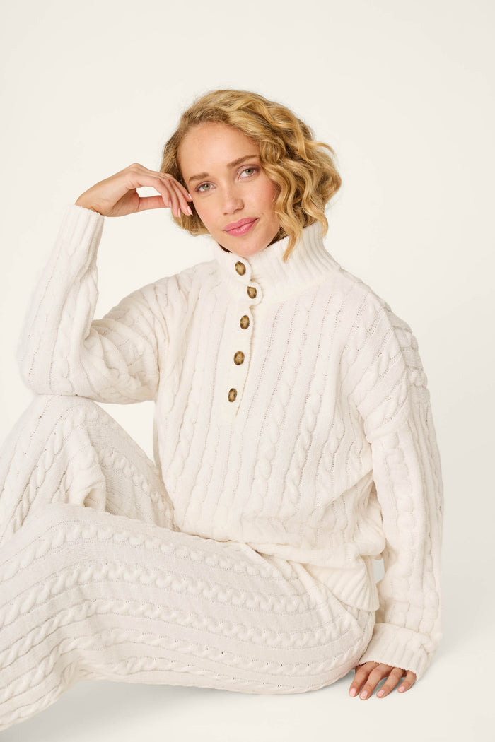A woman with short blond curly hair, dressed in the P.J. Salvage Cozy Chenille Cable Knit Ivory Pullover Lounge Set, sits with her head resting on her hand and smiles gently at the camera against a plain light background.