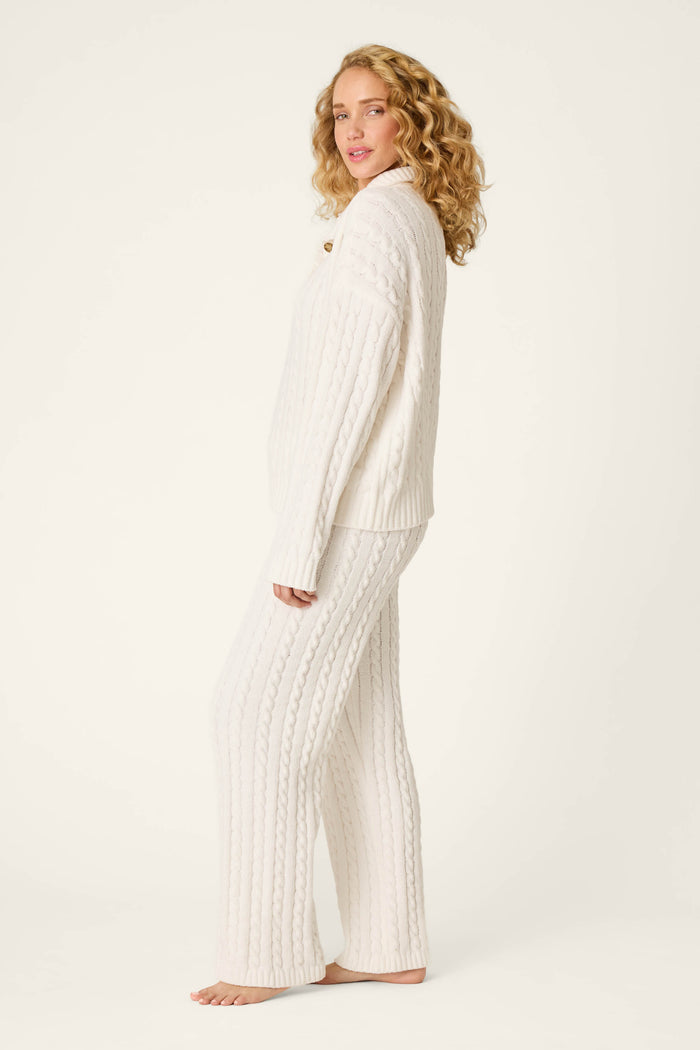 A woman with curly blonde hair models the Cozy Chenille Cable Knit Ivory Pullover Lounge Set by P.J. Salvage. She stands barefoot against a light background, dressed in the comfy off-white sweater and matching pants, gazing at the camera with a relaxed expression.