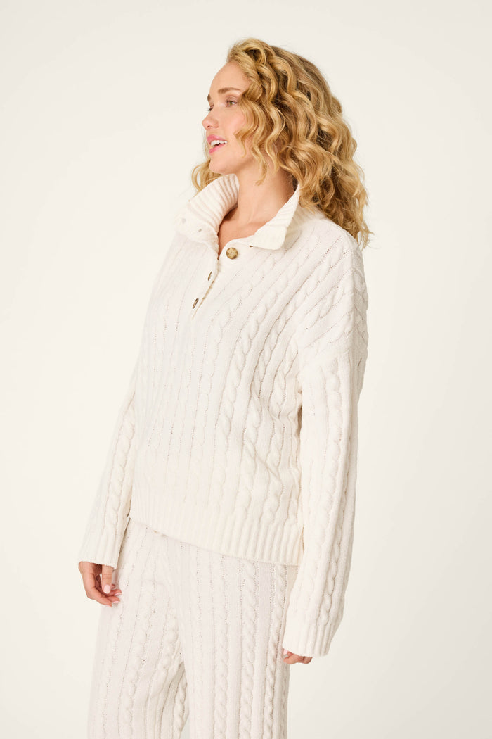 A woman with curly blonde hair wears the P.J. Salvage Cozy Chenille Cable Knit Ivory Pullover, standing against a plain light background, looking to the side and smiling.