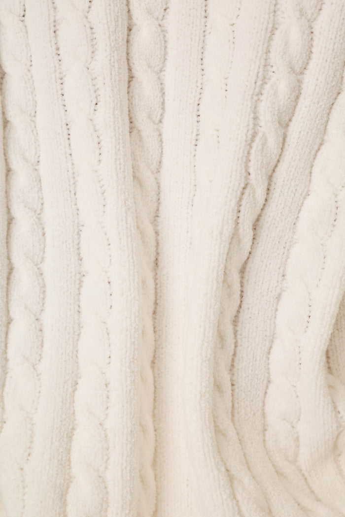 Close-up of a cream-colored cable-knit fabric texture, showing the detailed knit pattern and soft folds.