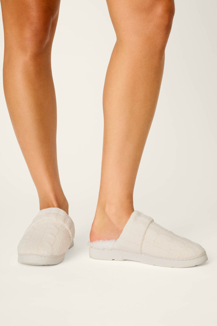 A person wears P.J. Salvage Cozy Chenille Cable Knit Ivory Slippers, featuring a soft textured knit design, while standing on a white background. Only their lower legs and feet are visible.