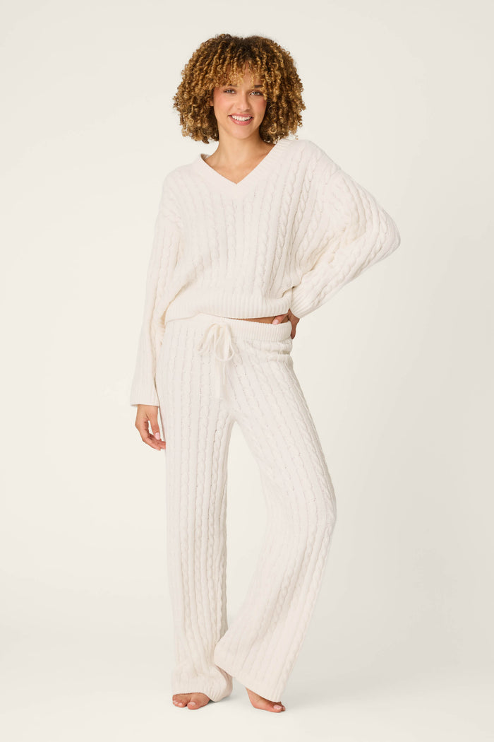 A person with curly hair wears the P.J. Salvage Cozy Chenille Cable Knit Ivory V-Neck Lounge Set, featuring a soft, cable-knit sweater and matching wide-leg pants. They stand barefoot, smiling with one hand on their hip against a plain white background.