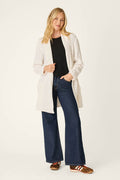 A person stands smiling against a plain light background, wearing the P.J. Salvage Cozy Chenille Cable Knit Long Ivory Cardigan over a black shirt, paired with blue wide-leg jeans and patterned slip-on shoes.