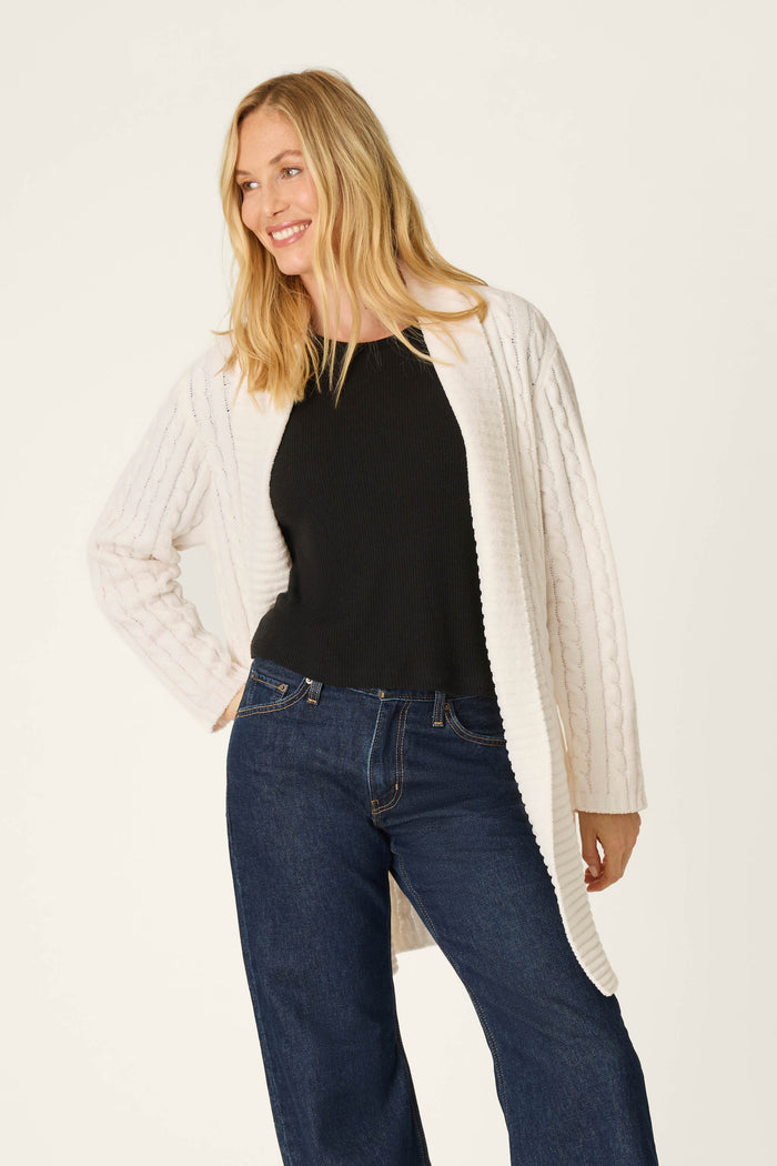 A woman with long blonde hair wears the P.J. Salvage Cozy Chenille Cable Knit Long Ivory Cardigan over a black top and blue jeans, smiling and looking to the side against a plain background.