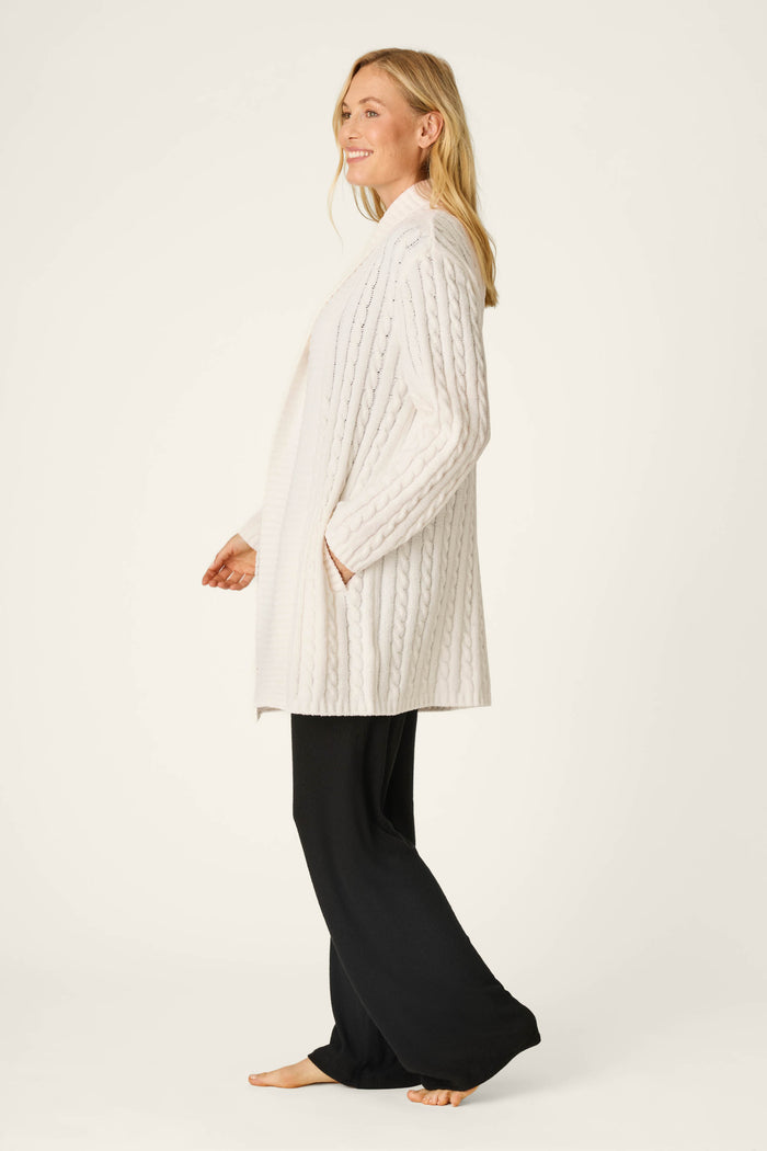 A woman stands barefoot, smiling in profile, wearing the P.J. Salvage Cozy Chenille Cable Knit Long Ivory Cardigan with pockets and loose black pants against a plain light background.