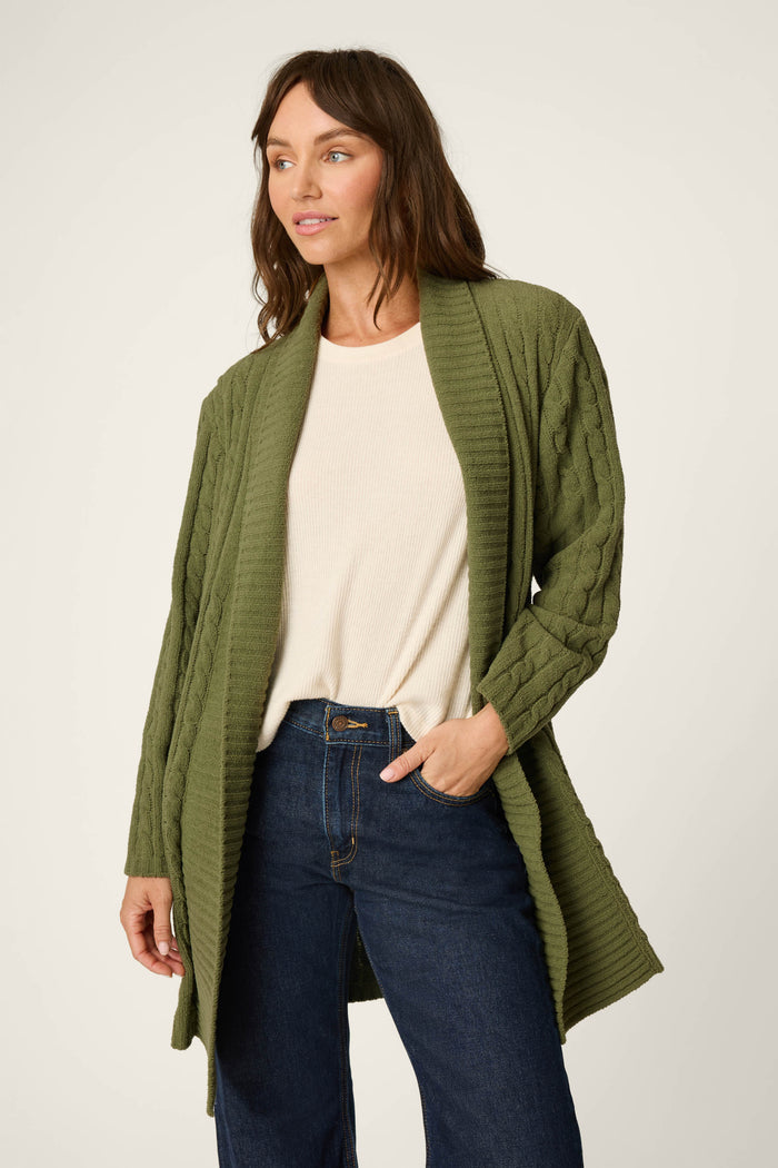A woman stands with one hand in her pocket, gazing slightly to the side against a plain, light background, wearing the P.J. Salvage Cozy Chenille Cable Knit Long Olivine Cardigan over a cream top and blue jeans.
