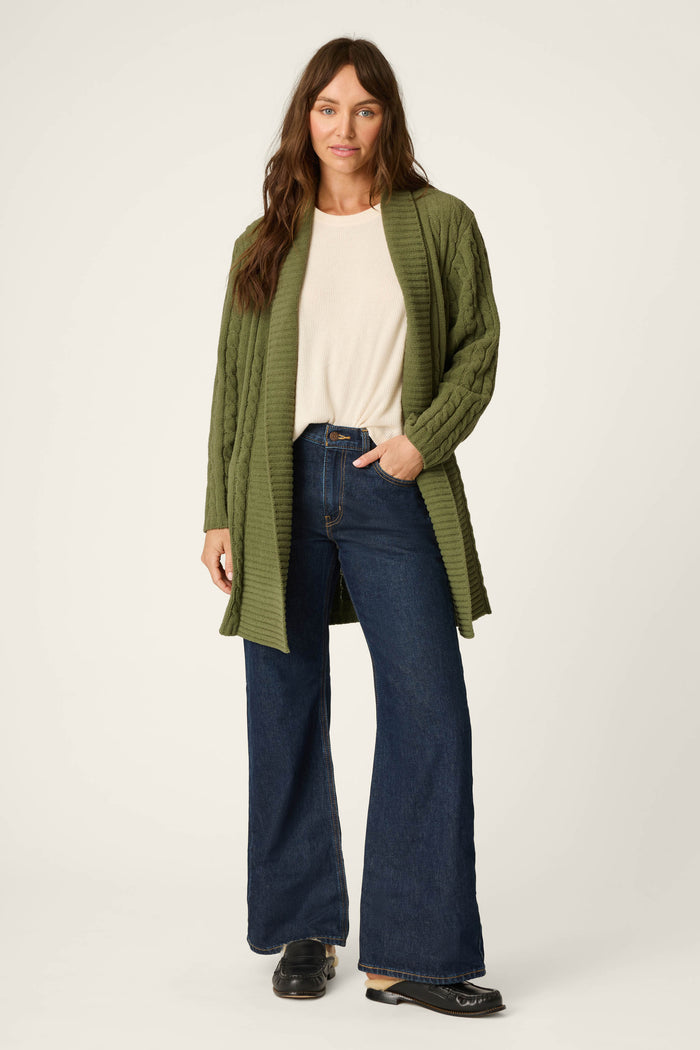 Wearing the P.J. Salvage Cozy Chenille Cable Knit Long Olivine Cardigan, a woman with long brown hair smiles softly with one hand in her pocket, paired with a cream top, dark blue wide-leg jeans, and black shoes as she stands against a plain light background.