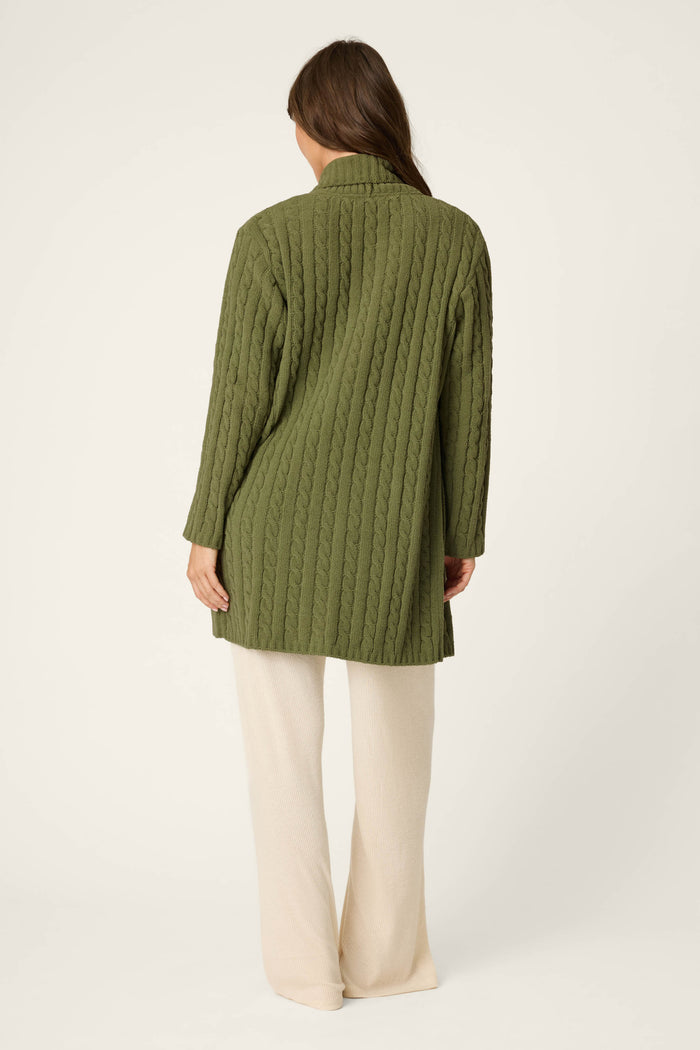 A woman with long brown hair stands facing away, wearing the P.J. Salvage Cozy Chenille Cable Knit Long Olivine Cardigan and cream wide-leg pants against a plain light background.