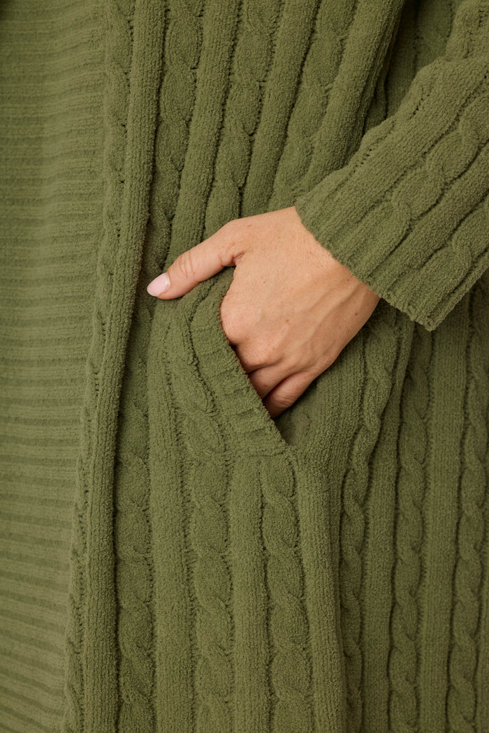 A close-up of a light-skinned hand resting inside the pocket of the Cozy Chenille Cable Knit Long Olivine Cardigan by P.J. Salvage.