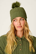 A woman with long blonde hair wears a green knit sweater and the P.J. Salvage Cozy Chenille Cable Knit Olivine Beanie with Pompom, looking slightly to the side.