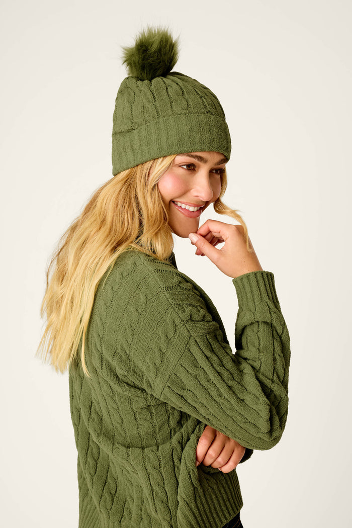 A woman smiles while looking to the side against a plain background, wearing the P.J. Salvage Cozy Chenille Cable Knit Olivine Beanie with Pompom and a matching green sweater.