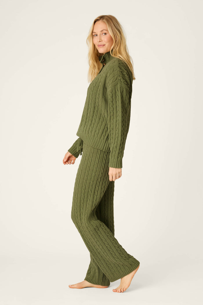 A woman with long blonde hair models the P.J. Salvage Cozy Chenille Cable Knit Olivine Pullover Lounge Set, featuring an olive green, cable-knit pullover and matching pants. She stands barefoot against a light background, gazing at the camera with a slight smile.