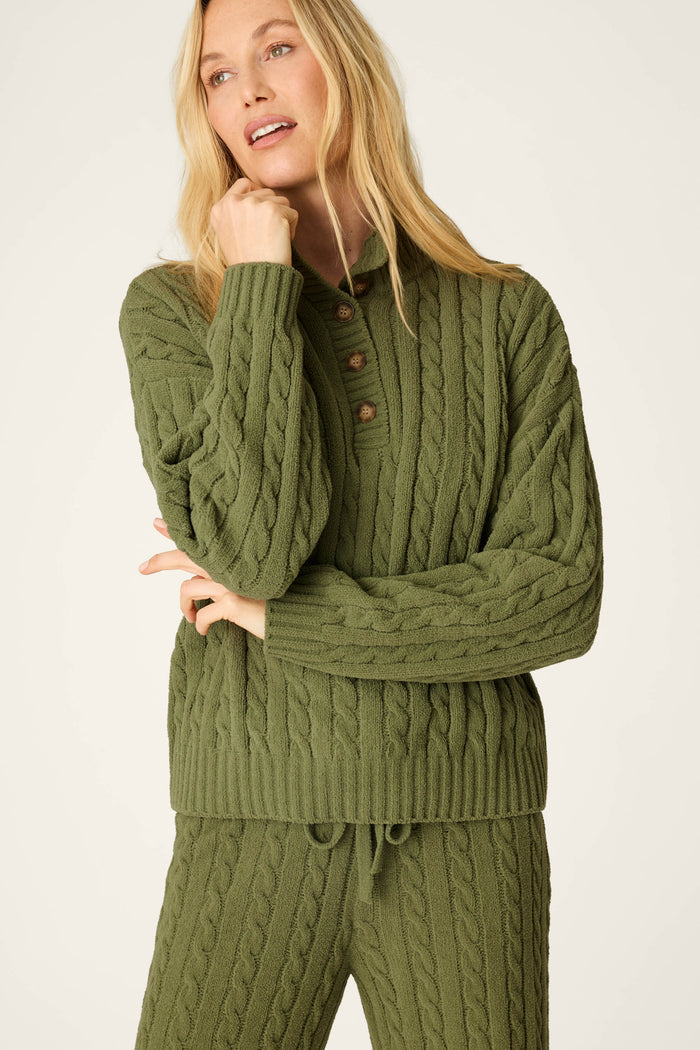 A woman models the P.J. Salvage Cozy Chenille Cable Knit Olivine Pullover Lounge Set, standing with one hand touching her chin and gazing off to the side.