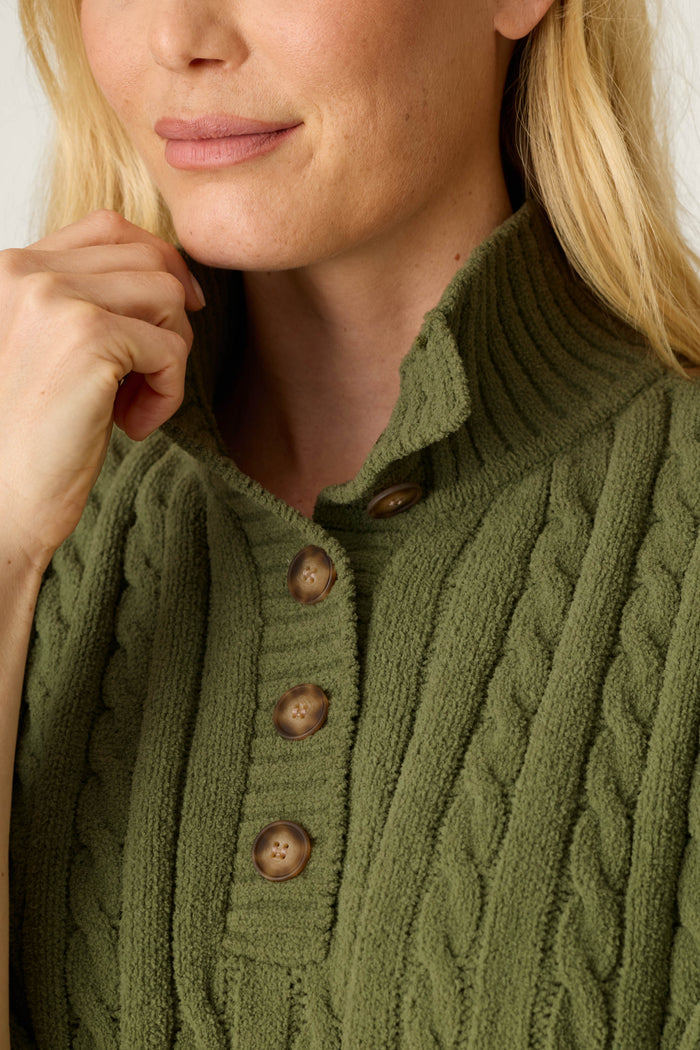 A woman with blonde hair is seen wearing the Cozy Chenille Cable Knit Olivine Pullover Lounge Set by P.J. Salvage, featuring a textured cable-knit design, brown buttons, and a high collar. She is holding the collar with her left hand, with only the lower part of her face and upper torso visible.